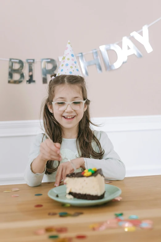 a little girl sitting at a table with a birthday cake, wearing black rimmed glasses, banner, grey, gen z