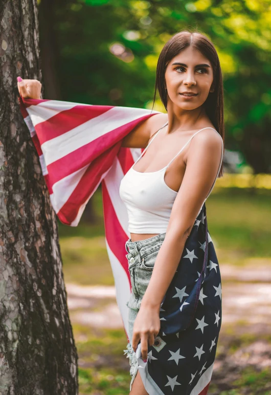 a woman standing next to a tree holding an american flag, wearing a tank top and shorts, mia khalifa, 2019 trending photo, 5 0 0 px models