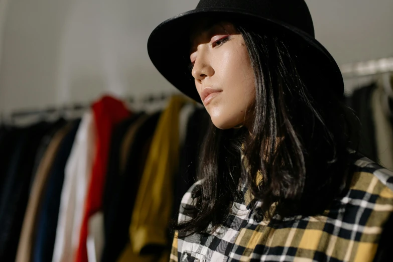 a woman standing in front of a rack of clothes, trending on pexels, visual art, wearing cylinder hat, sui ishida with black hair, flannel, demna gvasalia