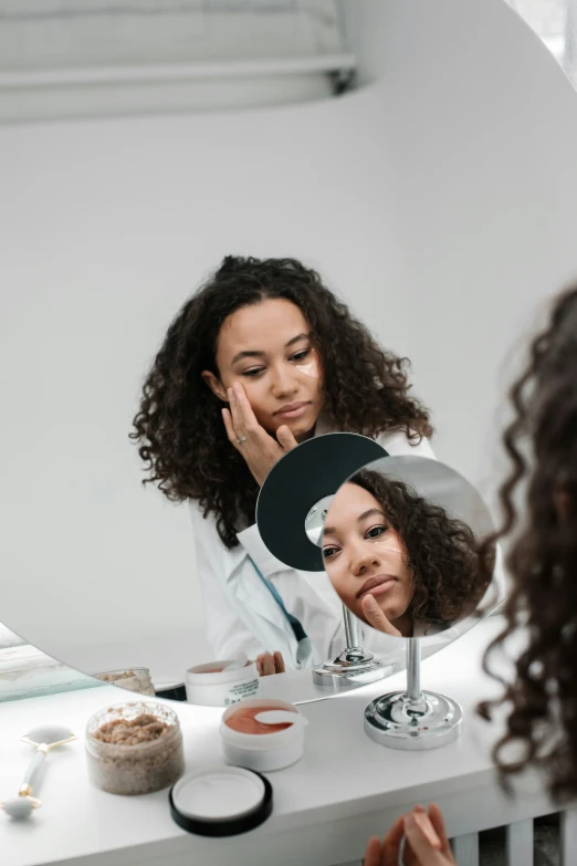 a woman getting her make - up done in front of a mirror, trending on pexels, happening, wearing a white lab coat, curly haired, set against a white background, facial scar