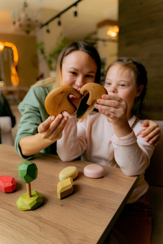 two girls sitting at a table eating doughnuts, by Julia Pishtar, pexels contest winner, wooden art toys, solid coloured shapes, the woman holds more toys, on wooden table