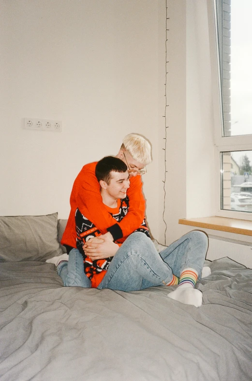 a man sitting on top of a bed next to a woman, by Zofia Stryjenska, trending on pexels, lgbt, wearing a sweater, two men hugging, orange jacket