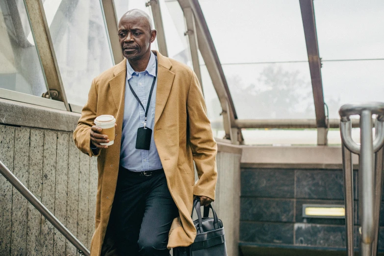 a man walking down a flight of stairs holding a cup of coffee, professor clothes, emmanuel shiru, light brown coat, holding a gold bag