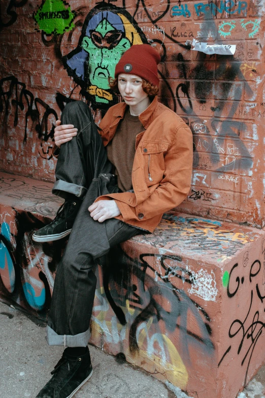 a man sitting on a bench in front of a graffiti covered wall, an album cover, inspired by Seb McKinnon, trending on pexels, brown pants, red haired teen boy, dressed in a ((ragged)), profile picture 1024px