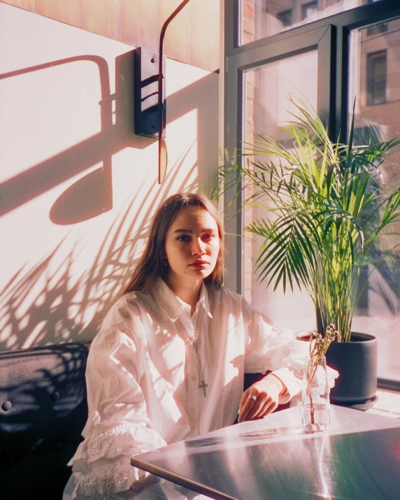 a woman sitting at a table in front of a window, a polaroid photo, inspired by Elsa Bleda, trending on unsplash, light and space, wearing a white button up shirt, next to a plant, portrait sophie mudd, sun behind him