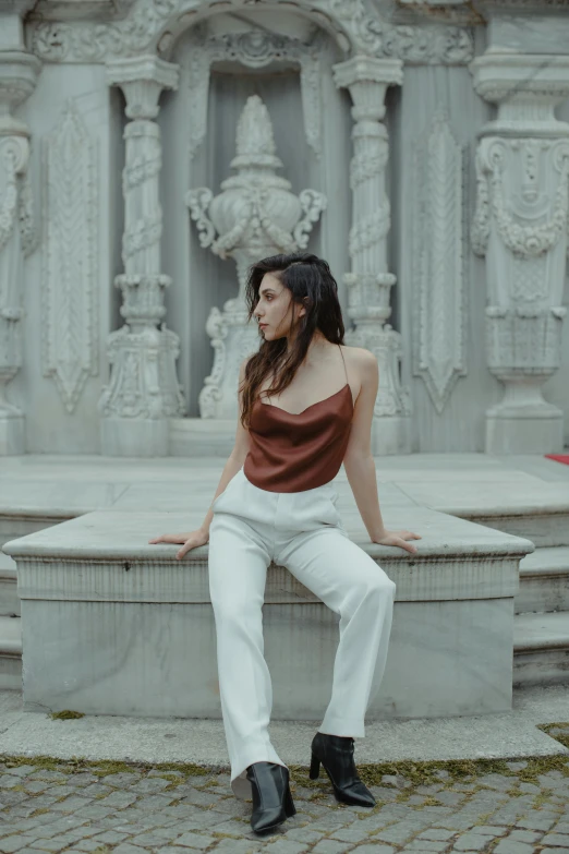 a woman sitting on a stone bench in front of a fountain, an album cover, inspired by Elsa Bleda, trending on pexels, renaissance, white pants, black and red silk clothing, white marble buildings, brown pants