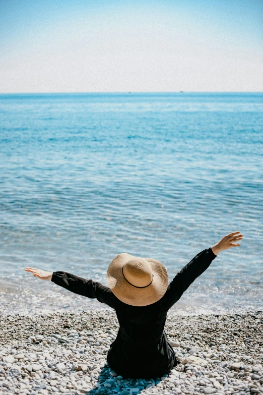 a woman sitting on top of a beach next to the ocean, pexels contest winner, renaissance, with arms up, black sun hat, happy italian beach scene, his arms spread. ready to fly