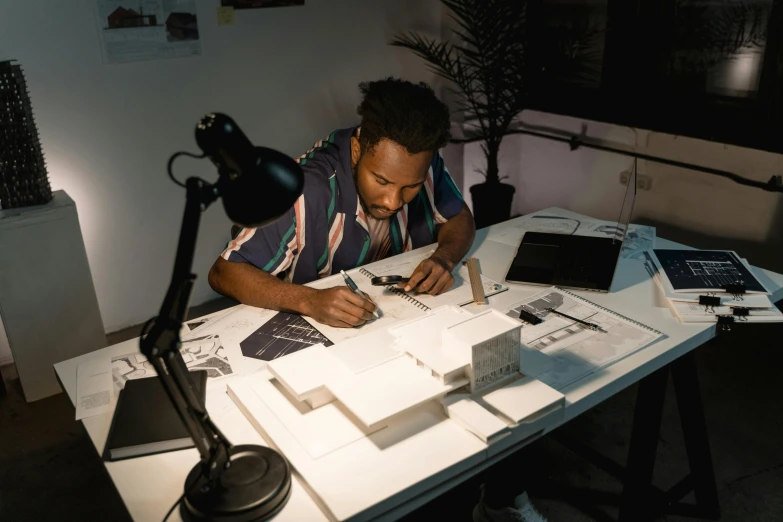 a man sitting at a desk writing on a piece of paper, a drawing, inspired by Afewerk Tekle, pexels contest winner, architectural model, avatar image, low lighting, artist wearing overalls