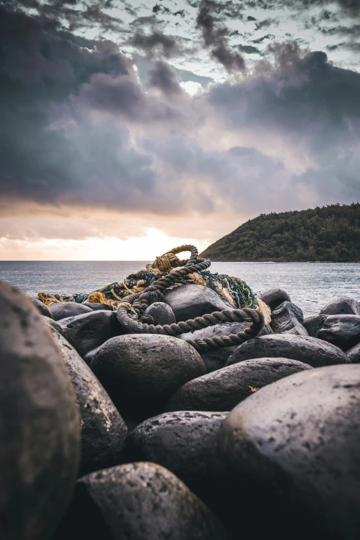 a rocky beach next to the ocean under a cloudy sky, by Jesper Knudsen, unsplash contest winner, ropes and chains, on an island, ((rocks)), brown cobble stones