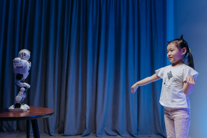 a little girl standing in front of a robot, by Jaakko Mattila, unsplash, footage of a theater stage, throwing cards in the air, blue backdrop, 15081959 21121991 01012000 4k