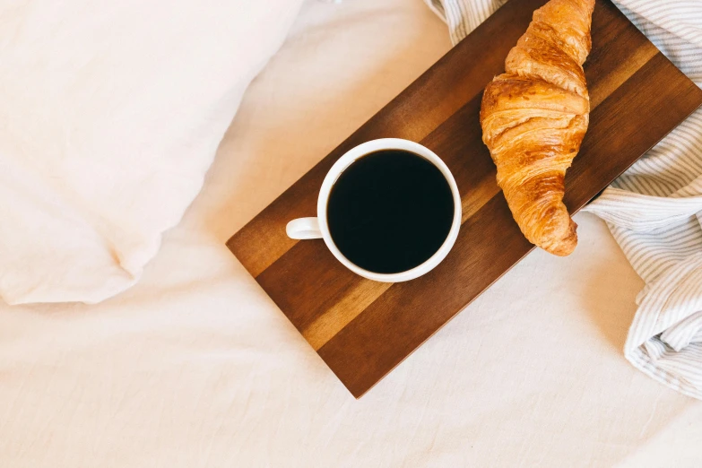 a wooden tray topped with a croissant next to a cup of coffee, trending on unsplash, romanticism, wearing a black robe, laying in bed, thumbnail, background image