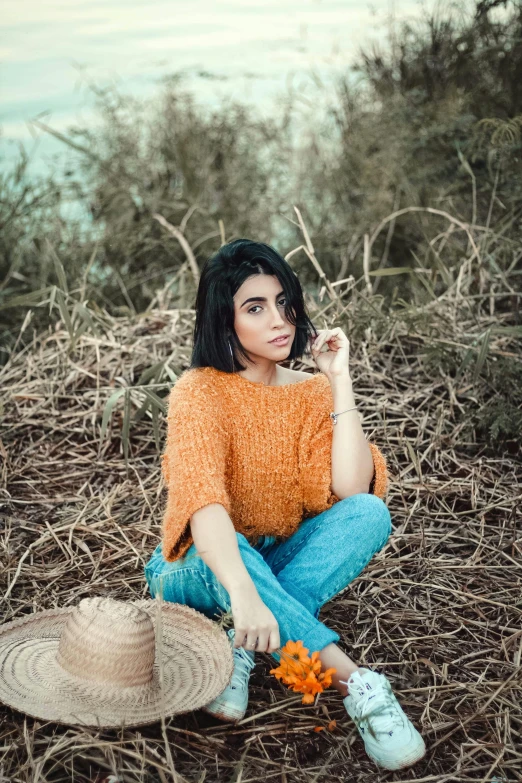 a woman sitting on the ground with a straw hat, a picture, inspired by Elsa Bleda, trending on pexels, hurufiyya, young middle eastern woman, wearing casual sweater, charli xcx, orange color