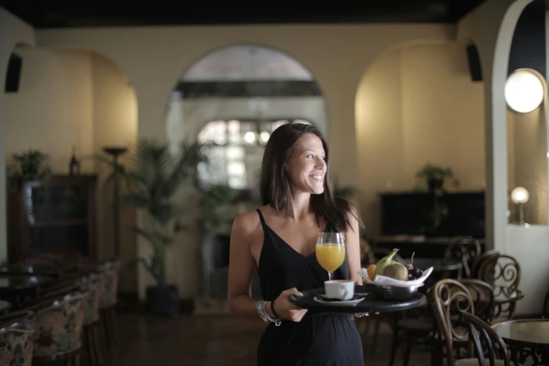 a woman in a black dress holding a tray of food, breakfast at las pozas, lounge, portrait image