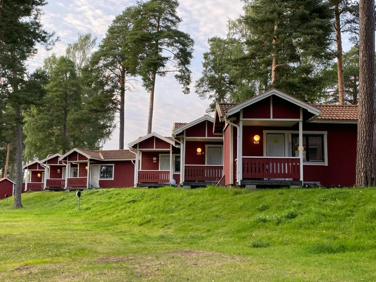 a row of red cabins sitting on top of a lush green hillside, by Jesper Knudsen, hurufiyya, pine trees in the background, exterior photo, front side, summer camp