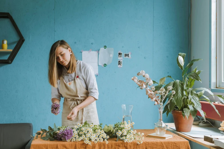 a woman standing in front of a table with flowers, a still life, trending on unsplash, process art, artist wearing overalls, filling the frame, blue and purple scheme, jasmine