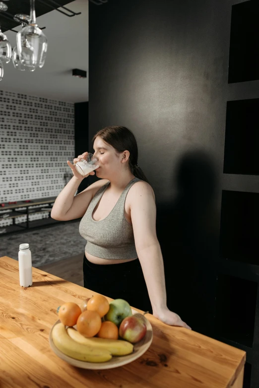 a woman drinking from a cup next to a bowl of fruit, she is wearing a black tank top, sport bra and shirt, profile image, kitchen counter