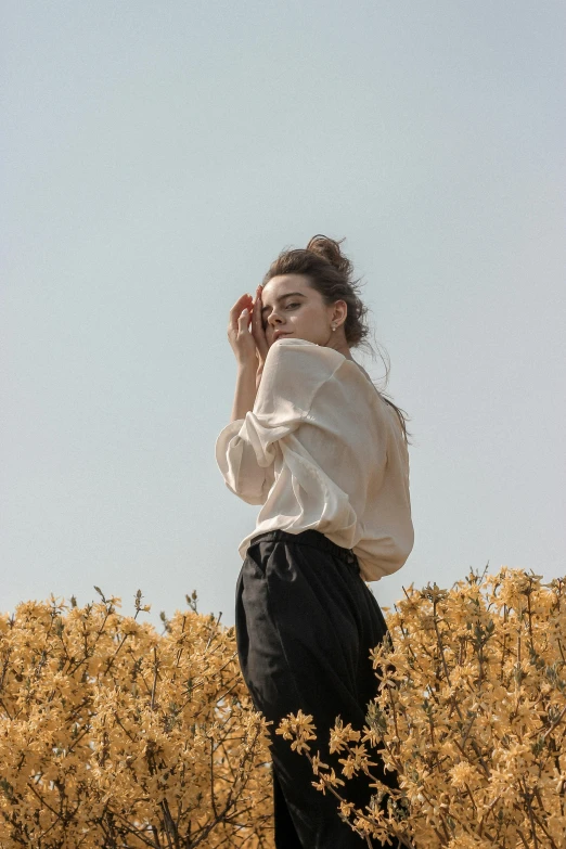 a woman standing in a field of yellow flowers, a picture, by irakli nadar, trending on unsplash, renaissance, wearing a linen shirt, pale beige sky, grim fashion model looking up, 3 / 4 pose