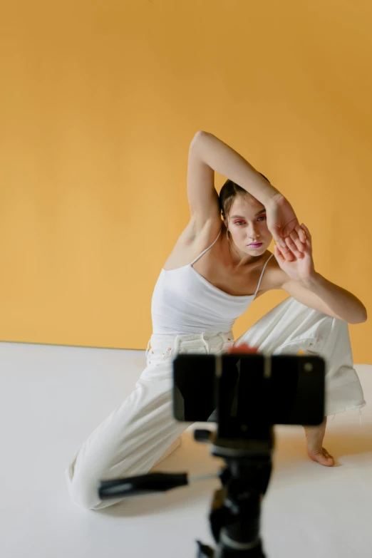 a woman sitting on top of a white floor next to a camera, inspired by Elizabeth Polunin, trending on pexels, video art, on a yellow canva, smartphone footage, dance meditation, face and upper - body focus