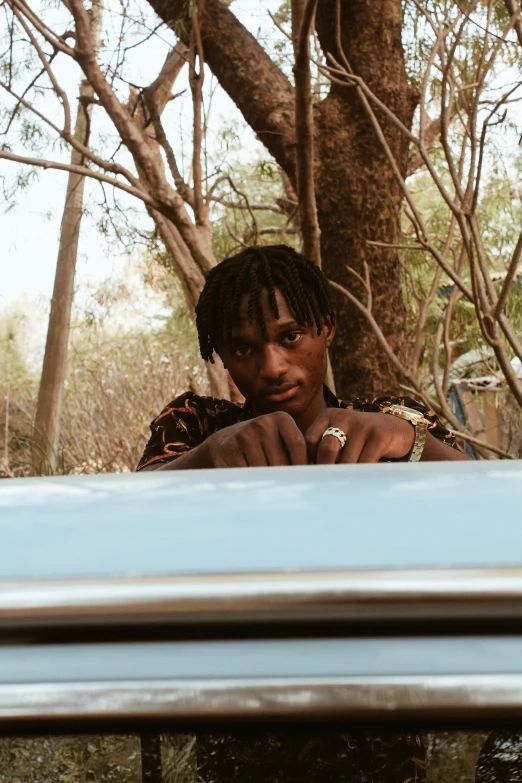 a man sitting on top of a car next to a tree, an album cover, by Maxwell Bates, trending on pexels, playboi carti, looks at the camera, ( ( theatrical ) ), in a jungle