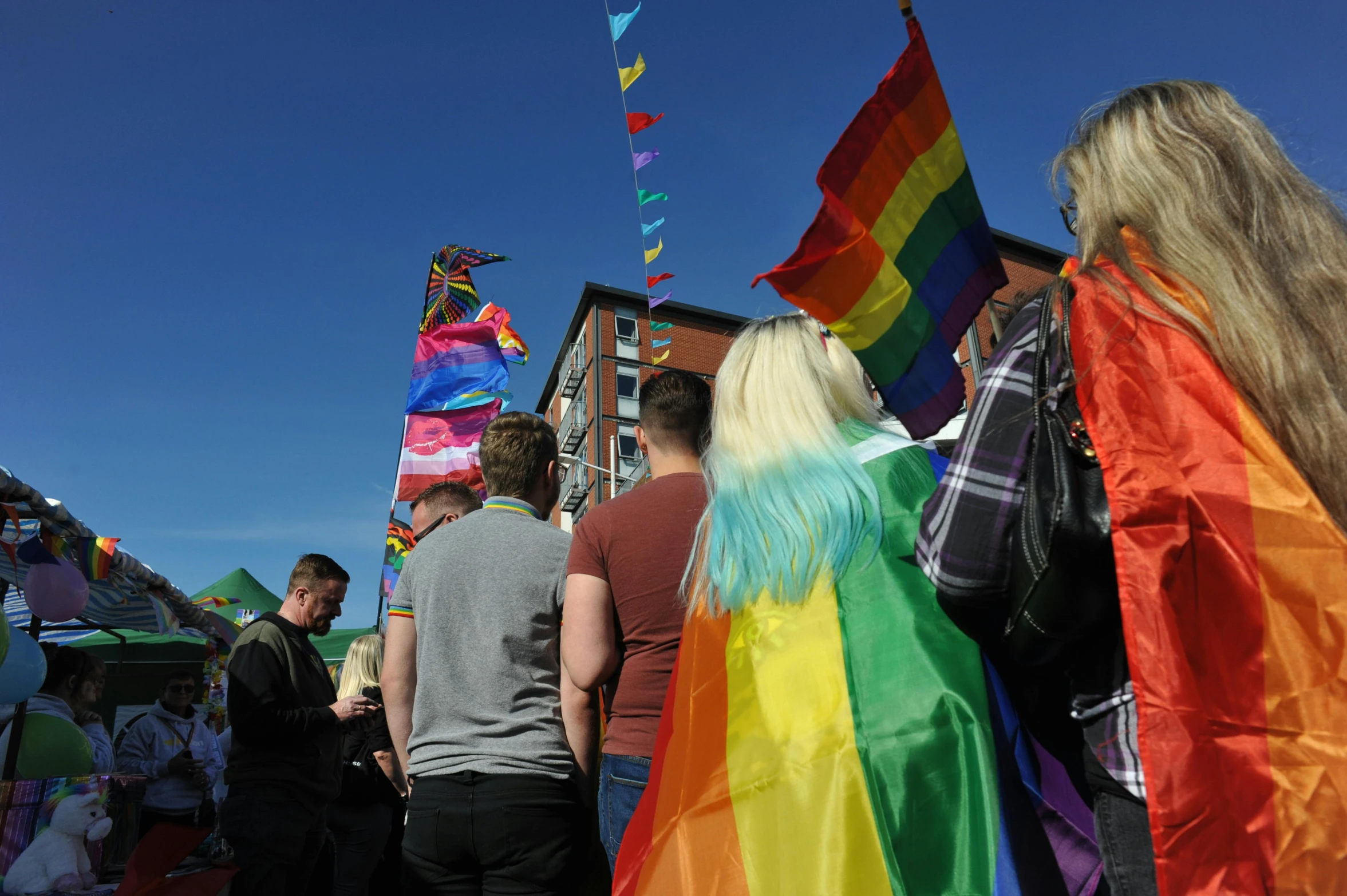 a group of people walking down a street holding flags, rainbow colours, nina tryggvadottir, blue sky, close-up shot taken from behind