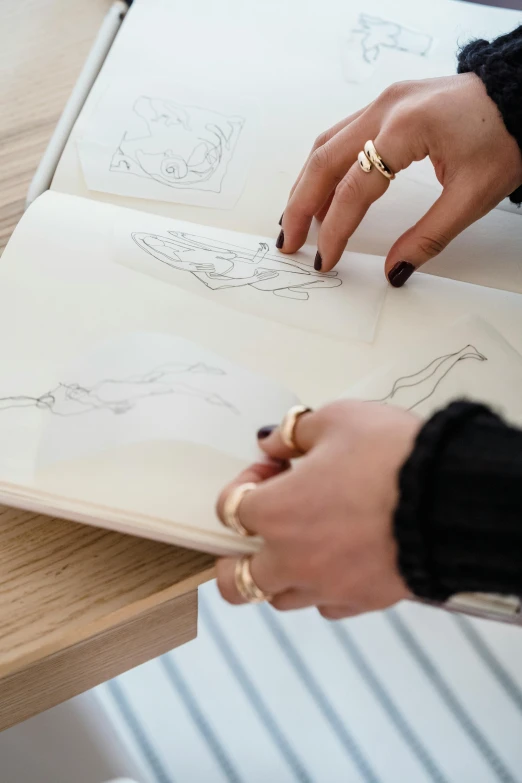 a woman sitting at a table drawing on a piece of paper, a drawing, by Nicolette Macnamara, trending on pexels, golden detailing, sketch book, sleek hands, in kaethe butcher
