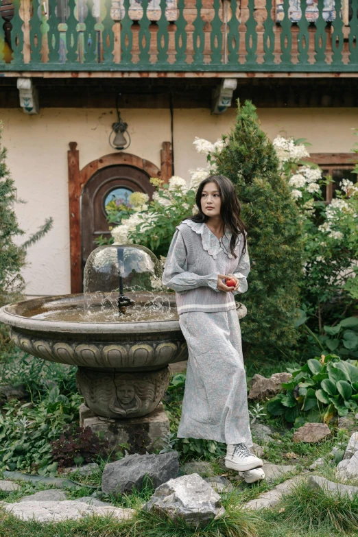 a woman standing next to a bird bath in a garden, a portrait, inspired by Li Di, unsplash, renaissance, gray hoodie, conde nast traveler photo, song hye - kyo, architectural digest