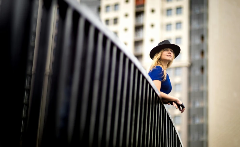 a woman in a hat leaning on a fence, by Daniel Gelon, perched on a skyscraper, square, taken with a canon eos 5d, a blond