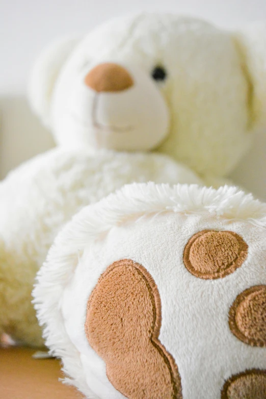 a white teddy bear sitting on top of a wooden table, white with chocolate brown spots, cushions, detail shot, twoddle
