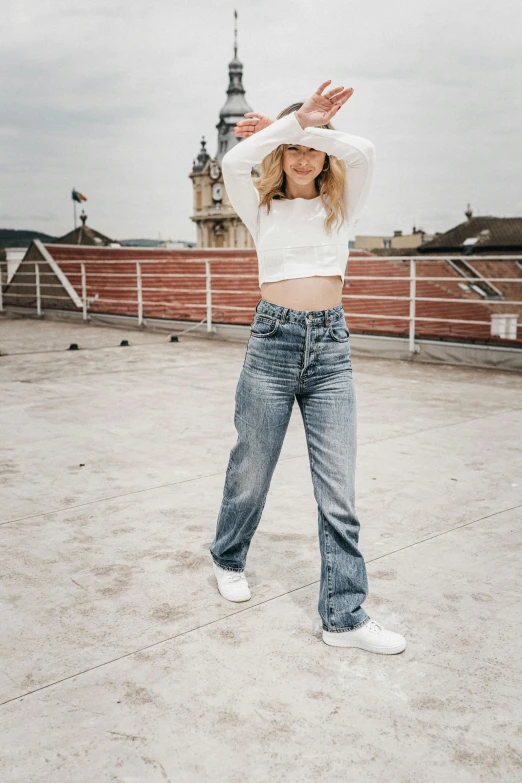 a woman standing on top of a roof with her hands in the air, an album cover, by Matija Jama, trending on pexels, renaissance, jeans pants, sydney sweeney, wearing white sneakers, midriff