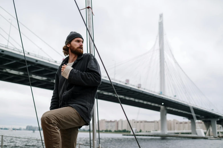 a man sitting on a boat in front of a bridge, a portrait, by Joseph Severn, pexels contest winner, wearing jacket, alexey egorov, standing on the mast, portrait of tall