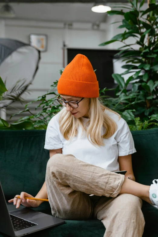 a woman sitting on a couch using a laptop, by Julia Pishtar, trending on pexels, beanie, orange roof, filled with plants, academic clothing