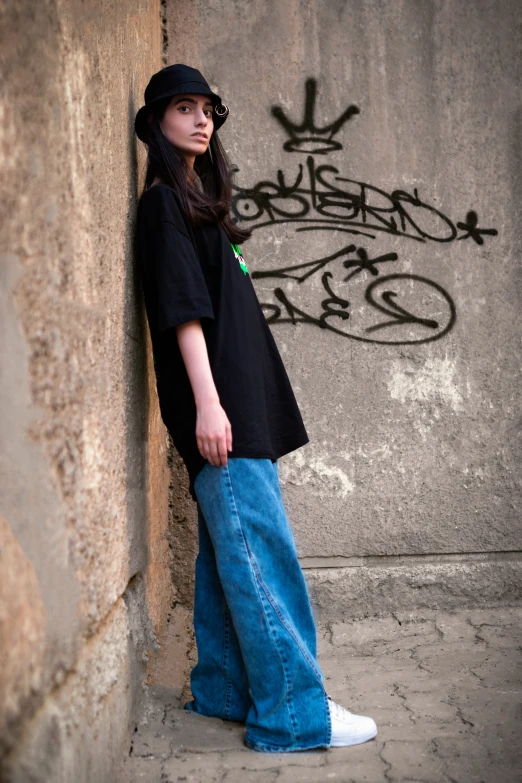 a woman leaning against a wall with graffiti on it, black t shirt, baggy jeans, official store photo, postprocessed)