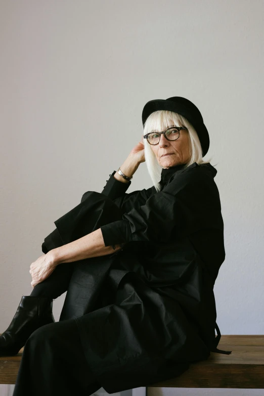 a woman sitting on top of a wooden bench, medium length slick white hair, wearing black rimmed glasses, in front of white back drop, wearing black dress and hat