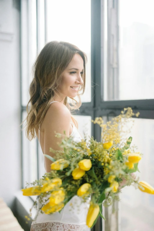a woman holding a bouquet of yellow flowers, camilla luddington, looking out window, in a white boho style studio, smiling
