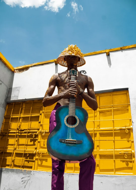 a man with a guitar standing in front of a building, inspired by Candido Bido, pexels contest winner, afrofuturism, in blue and yellow clothes, carnaval de barranquilla, die antwoord music video, wearing a straw hat and overalls