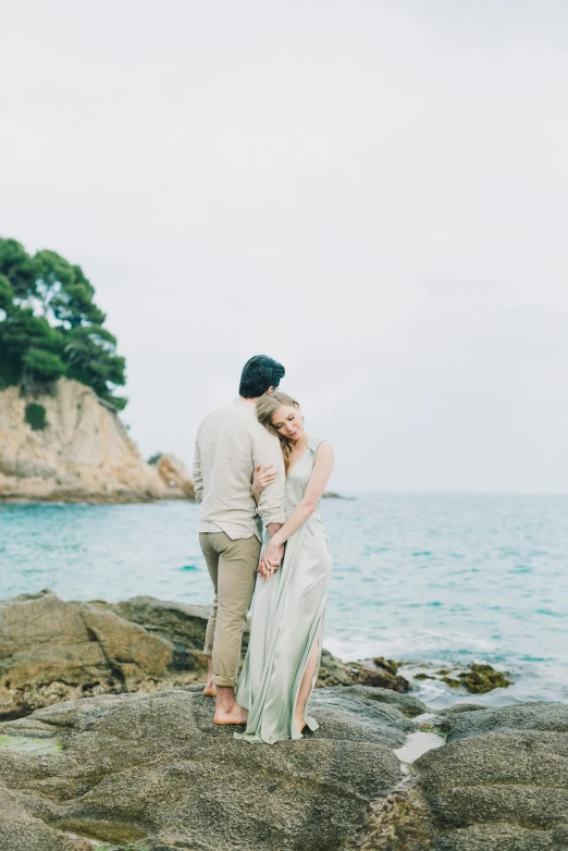 a man and woman standing on a rock next to the ocean, a picture, by Silvia Pelissero, unsplash, romanticism, 2 5 6 x 2 5 6 pixels, soft elegant gown, mediterranean, 15081959 21121991 01012000 4k