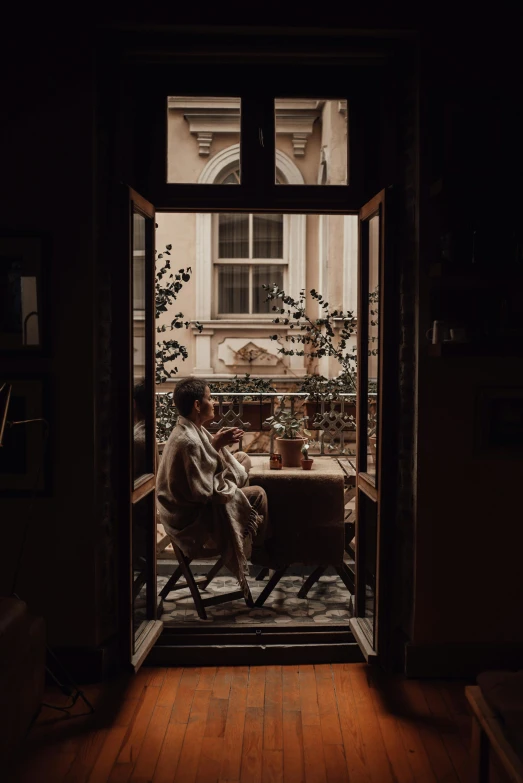 a person sitting in a chair looking out a window, inspired by Elsa Bleda, pexels contest winner, australian tonalism, wearing brown robes, balcony door, cozy cafe background, cinematic full body shot