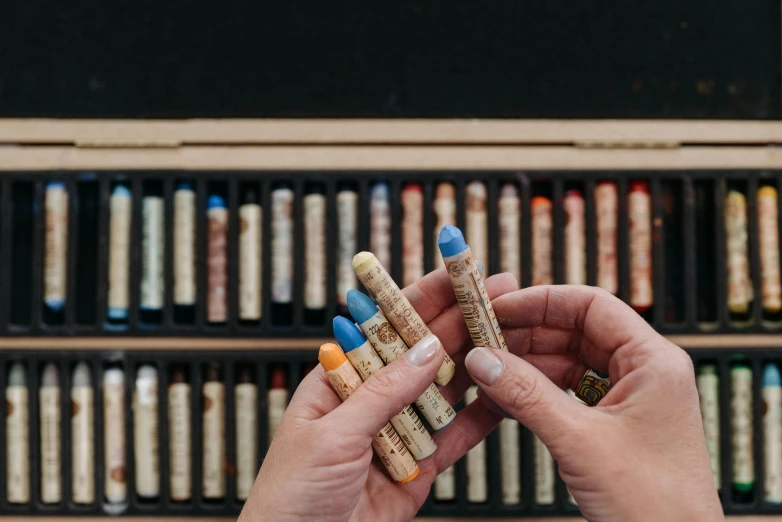 a person holding a bunch of crayons in front of a wall of crayons, by Sylvia Wishart, pexels contest winner, visual art, cigarrette boxes at the table, prussian blue and raw sienna, with intricate detail, black chalk