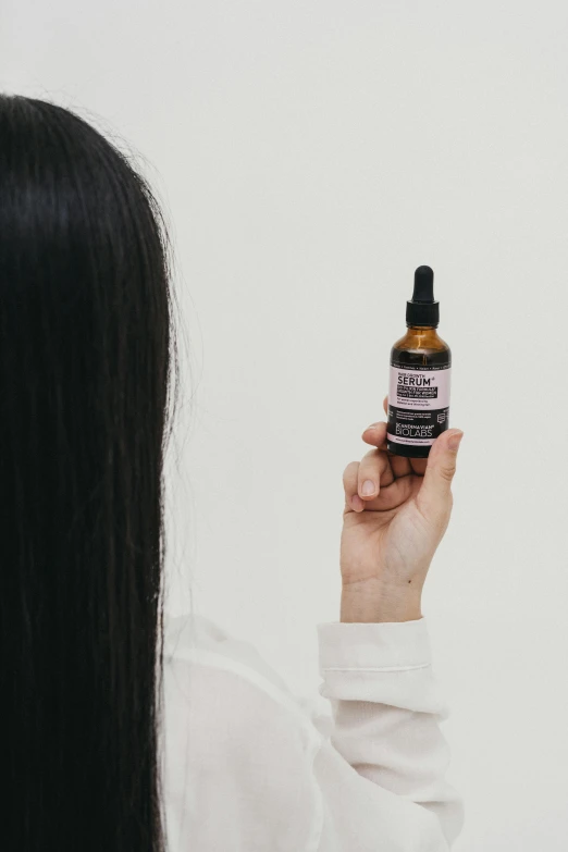a woman holding a bottle of hair oil, unsplash, in front of white back drop, back view also, apothecary, thick and bold black outlines