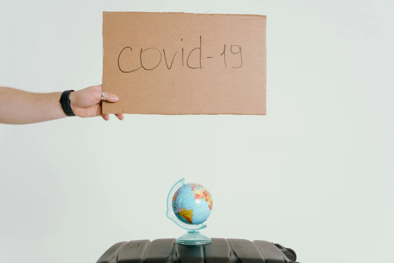 a person holding a sign that says covid 19, by Olivia Peguero, pexels contest winner, earth globe on top, in suitcase, made of cardboard, slightly turned to the right