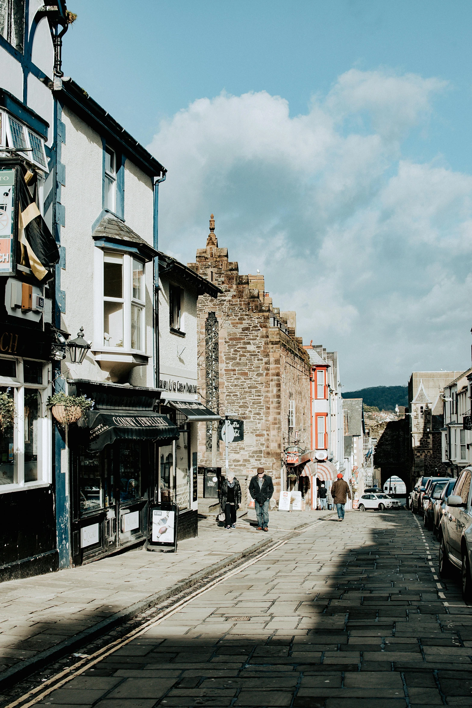 a group of people walking down a street next to tall buildings, medieval tumbledown houses, black mountains, quirky shops, an beautiful