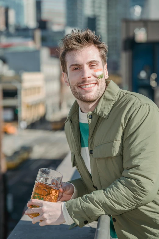 a man holding a glass of beer on top of a building, warm friendly face, wearing green jacket, tinder profile, city rooftop