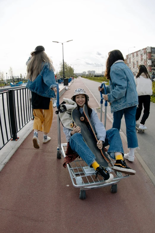 a group of people walking down a sidewalk with a skateboard in a cart, an album cover, by Attila Meszlenyi, unsplash, wearing a jeans jackets, young women, bridge, posing for the camera