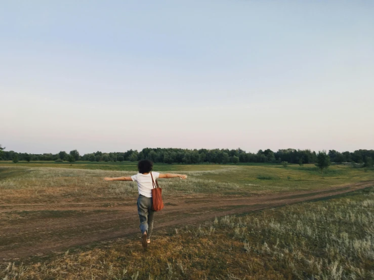 a person walking on a dirt road in a field, spreading her wings, jovana rikalo, looking onto the horizon, in a large grassy green field