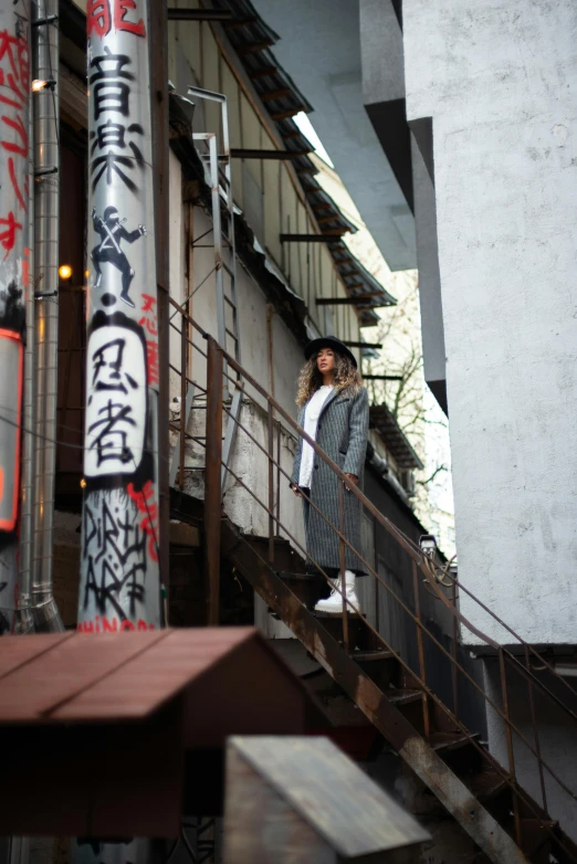 a man standing on the steps of a building, a picture, inspired by Okamoto Tarō, unsplash, woman is in a trenchcoat, graffiti in the background, like jiufen, seen from below