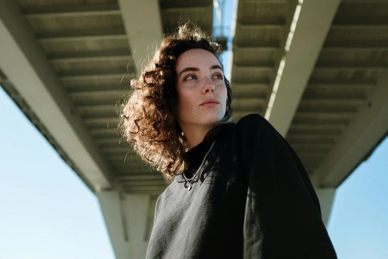 a woman with curly hair standing under a bridge, a portrait, by Winona Nelson, pexels contest winner, black sweater, flat colour, charli bowater, looking from shoulder