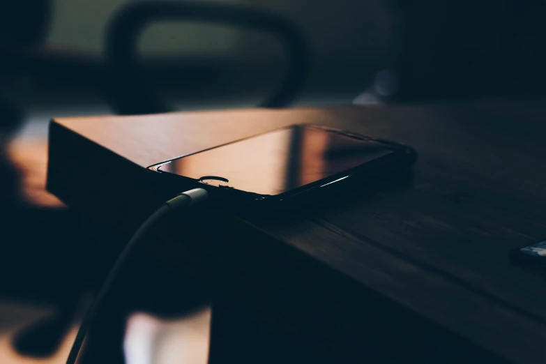 a cell phone sitting on top of a wooden table, by Niko Henrichon, trending on pexels, happening, cable plugged in, lo fi, dimly - lit, sitting on a desk