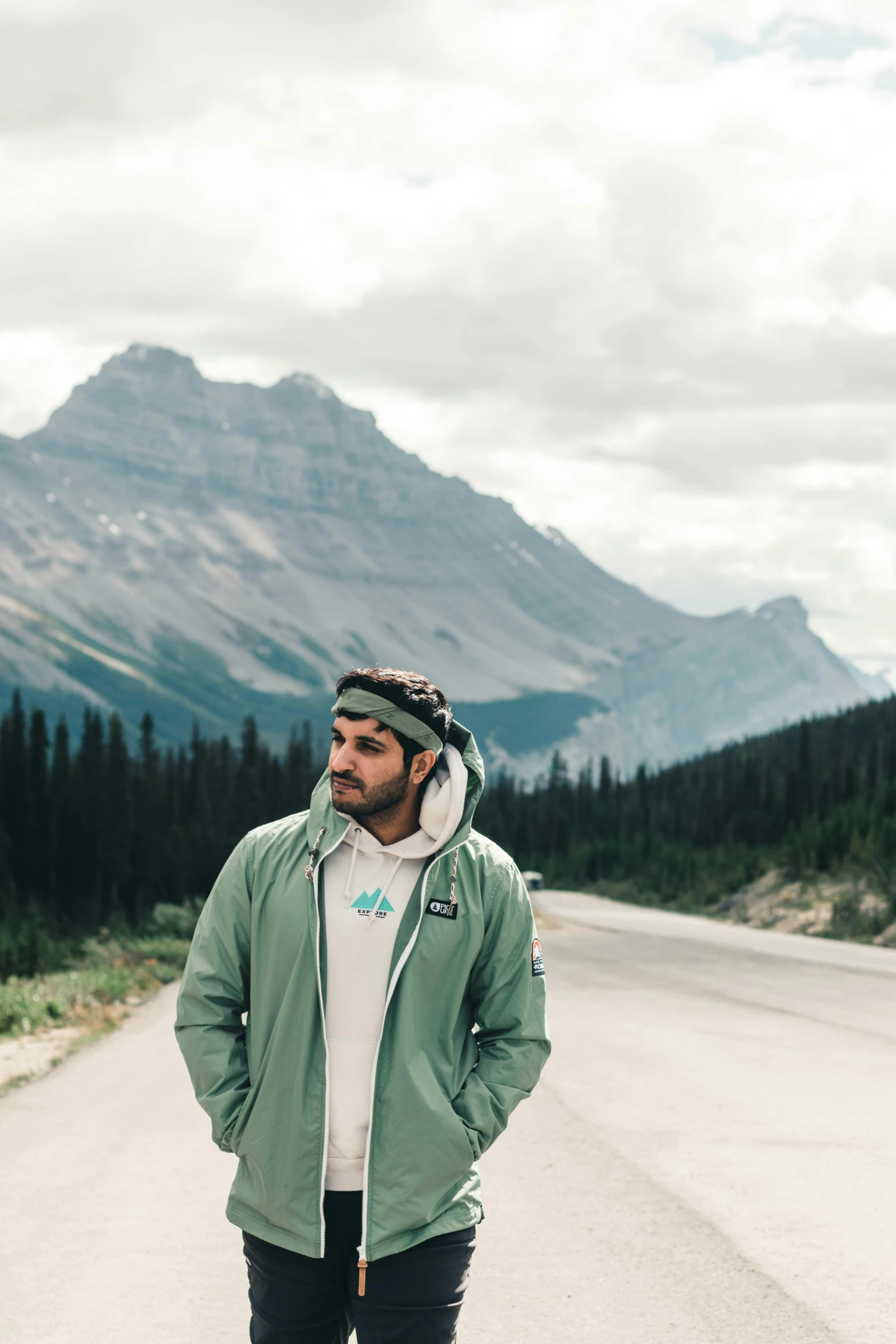 a man standing on the side of a road, a colorized photo, inspired by James Pittendrigh MacGillivray, trending on unsplash, green colored bomber jacket, banff national park, wearing a light grey crown, mountains in a background