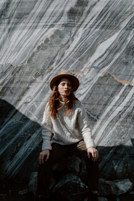 a woman sitting on top of a pile of rocks, inspired by Elsa Bleda, trending on pexels, renaissance, caracter with brown hat, on a large marble wall, wearing a white sweater, concert photo