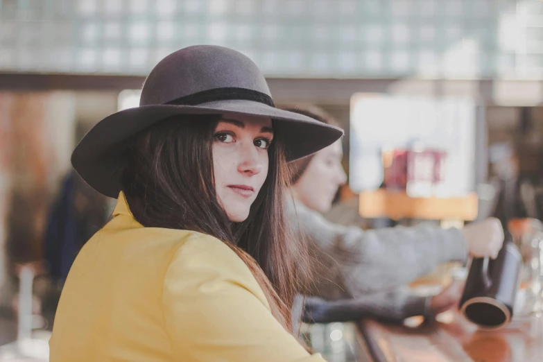 a woman sitting at a bar wearing a hat, by Emma Andijewska, pexels contest winner, renaissance, yellow and charcoal, avatar image, brunette, black stetson and coat